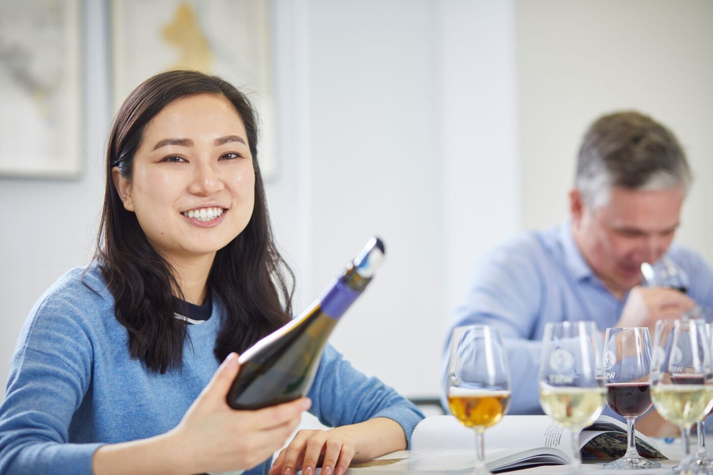 Woman smiling in the calendar while holding up a bottle of wine. Man in background with a wine glass at his nose.
