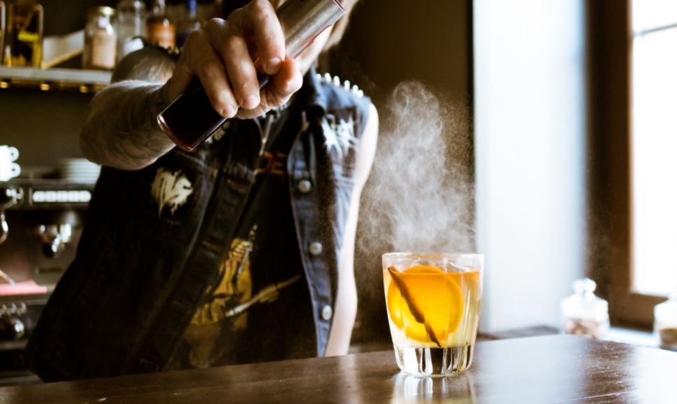Male bartender behind a bar pouring a liquid into a cocktail with an orange slice and a cinnamon stick