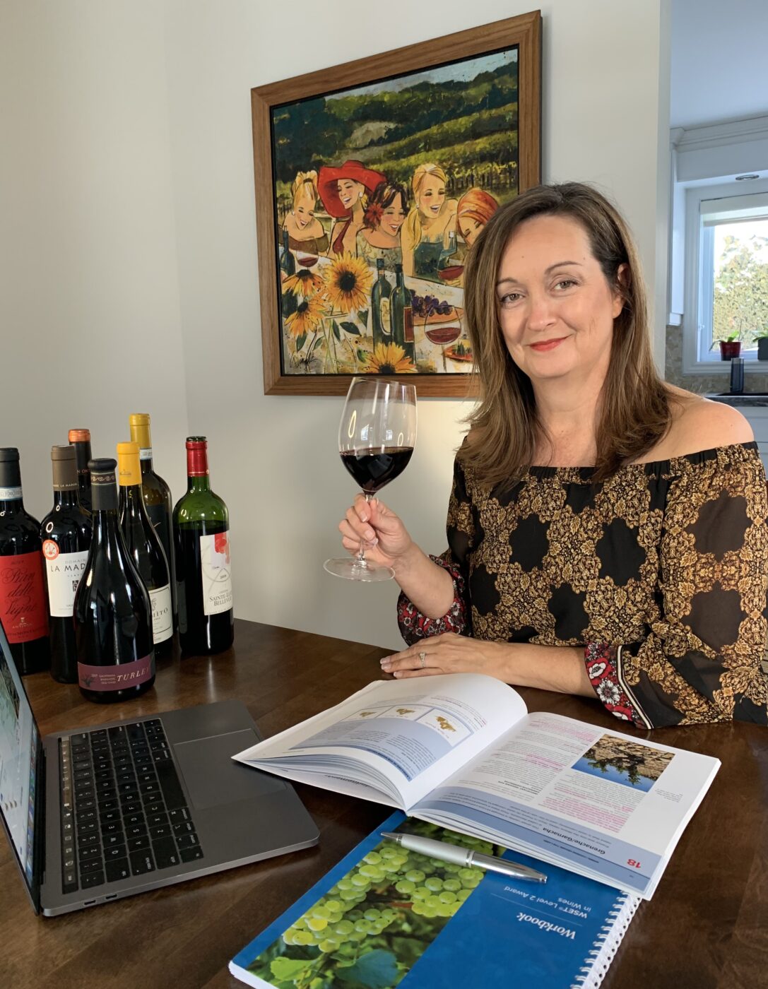 Woman sitting a table with bottles of wine and text books while holding a glass of red wine