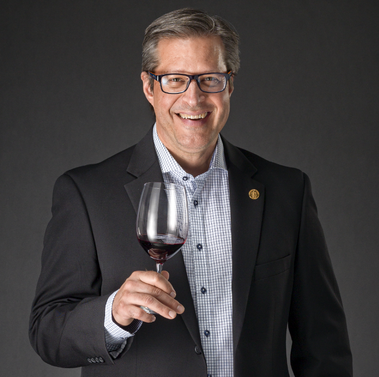 Man in a blazer and glasses smiling while holding up a glass of red wine