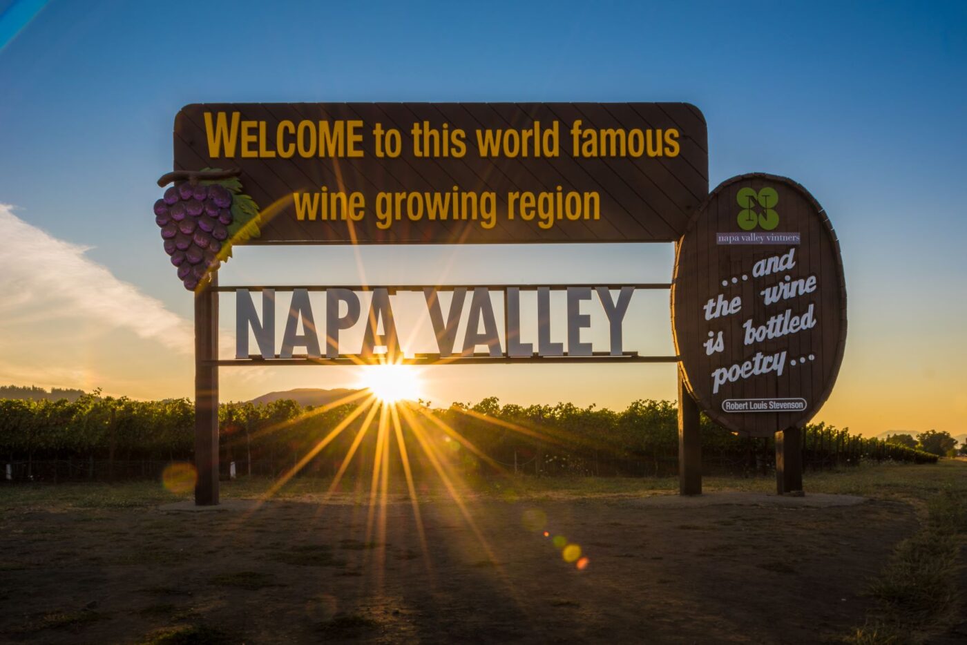 Image of a sign in front of a vineyard with the reflection of the sun. The sign says Napa Valley, welcome to this world famous wine growing region... and the wine is bottle poetry...
