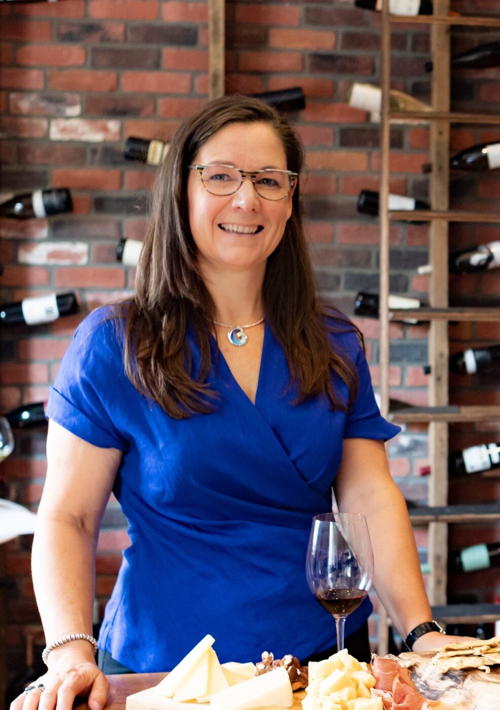 Woman in a blue shirt standing in a room with wine bottles on the wall in the background