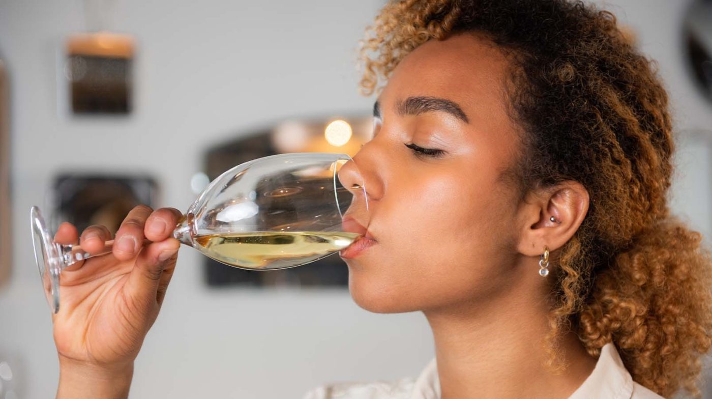 Side profile of a woman with curly hair and earrings tasting a glass of white wine.