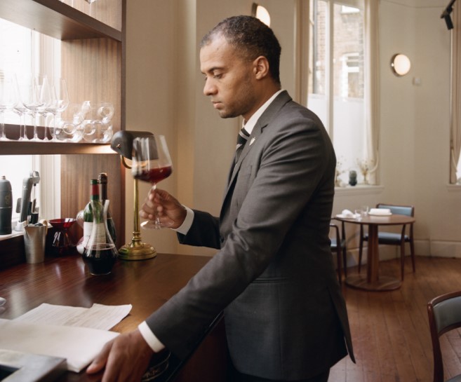 Man in a suit at a high table in a restaurant swirling a glass of red wine while looking at notes.