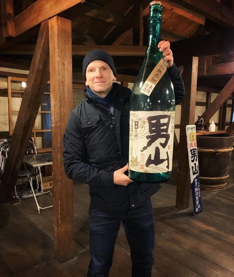 Man wearing a black hat smiling while holding up an extremely large bottle of sake.