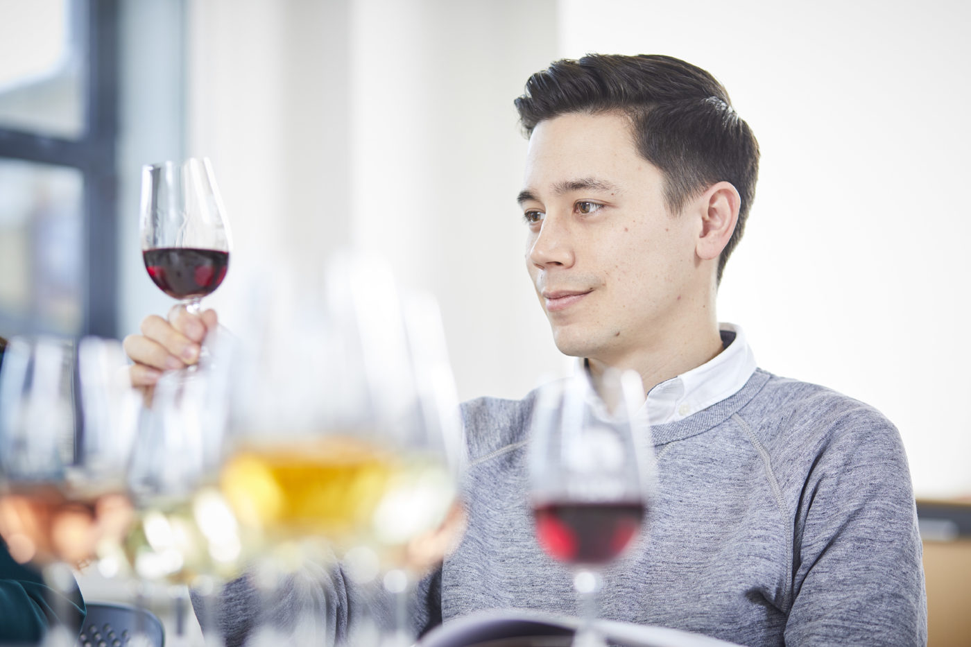 Man sitting at t table with various glasses of wine in front him. He is holding up and looking at a red wine.