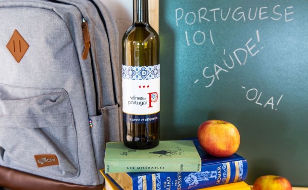 Bottle of wine on a pile of books with a backpack and blackboard in the background. The bottle says wines of Portugal. The blackboard says Portuguese 101, saude! and ola!