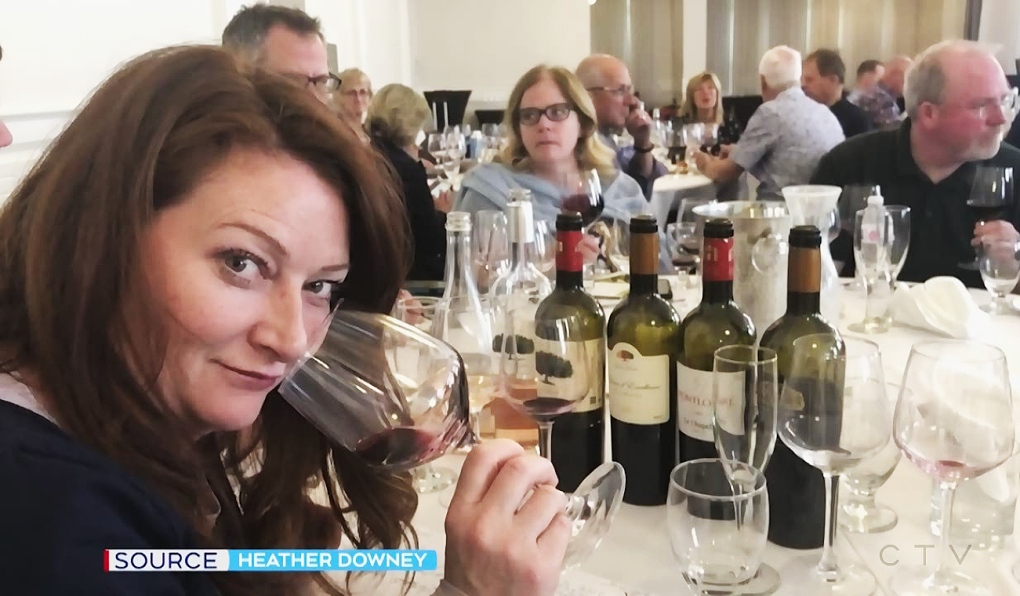 Woman with long brown hair looking into the camera while holding a glass of red wine to her nose. In front of her is a four bottles of wines and various glasses. Others are sitting ather table with various glasses as well.