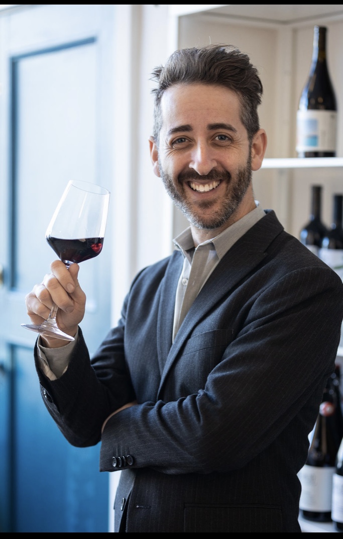 Man in a suit and beard in front of bottles on a shelf holding a glass of red wine and smiling with one arm crossed under the other.