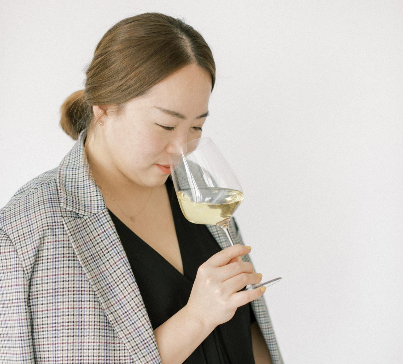 Woman with a grey plaid blazer over her shoullders looking down and smelling a glass of white wine