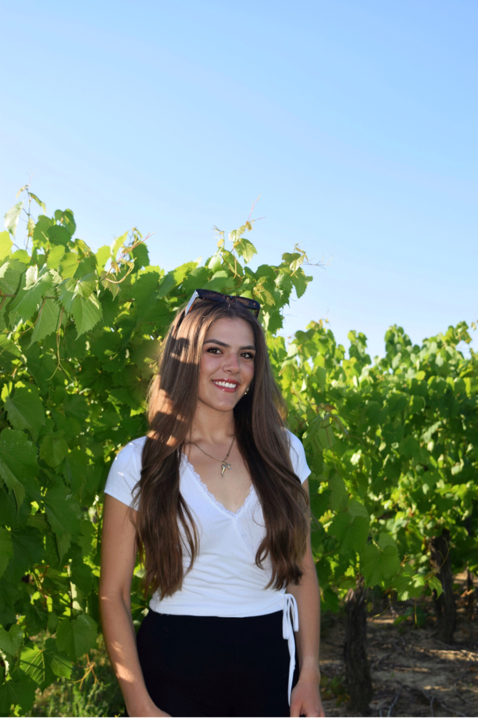 Woman with long brown hair in a white t-shirt smiling while standing in a vineyard