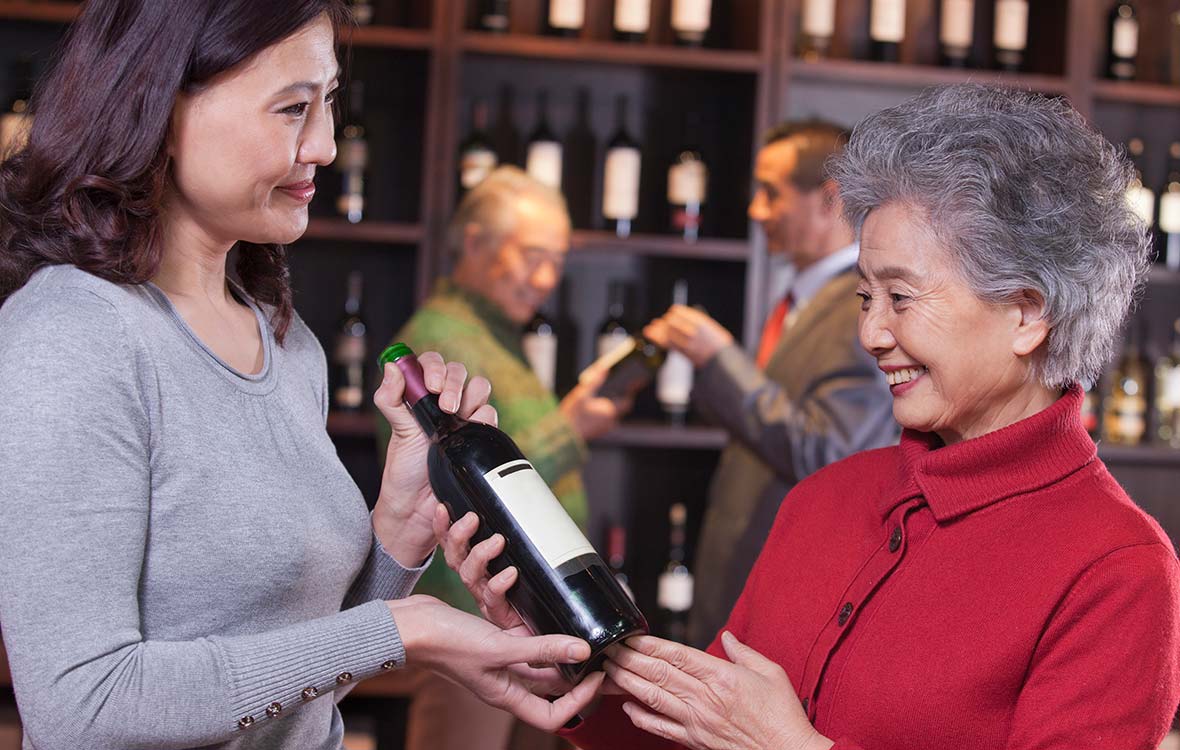 A younger woman hands a bottle of wine while smiling to an older woman who is also smiling.