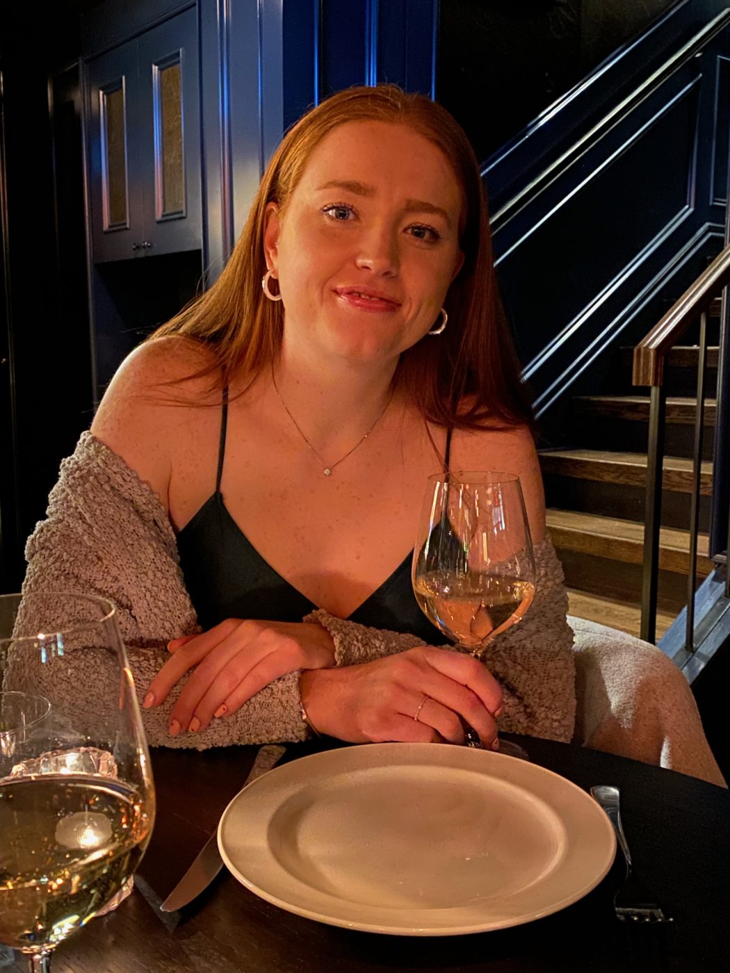 A young woman in a black dress and grey shawl sitting at a dining table holding a glass of white wine.