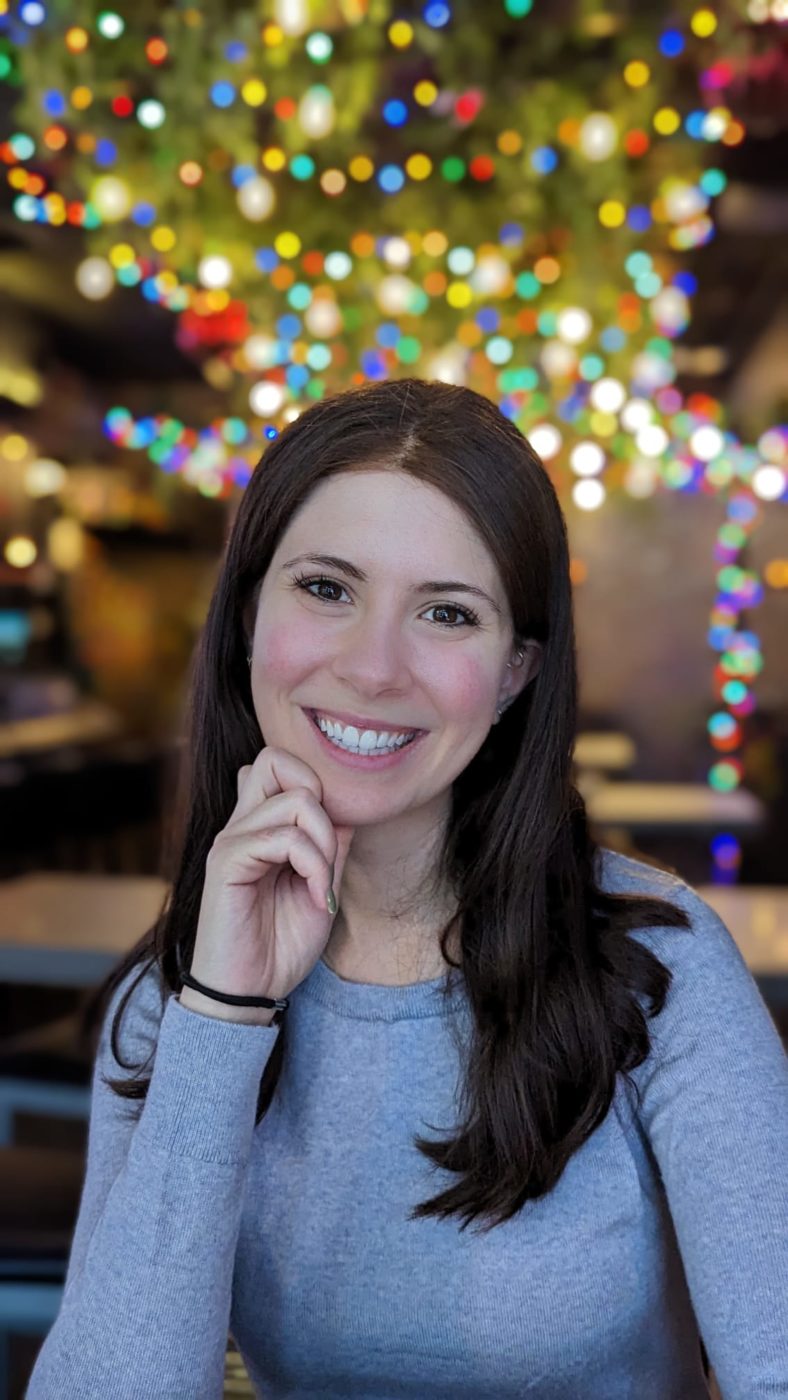 Woman with brown hair smiling with her hand to her face. She is wearing a light blue top and there are colourful lights in the background.