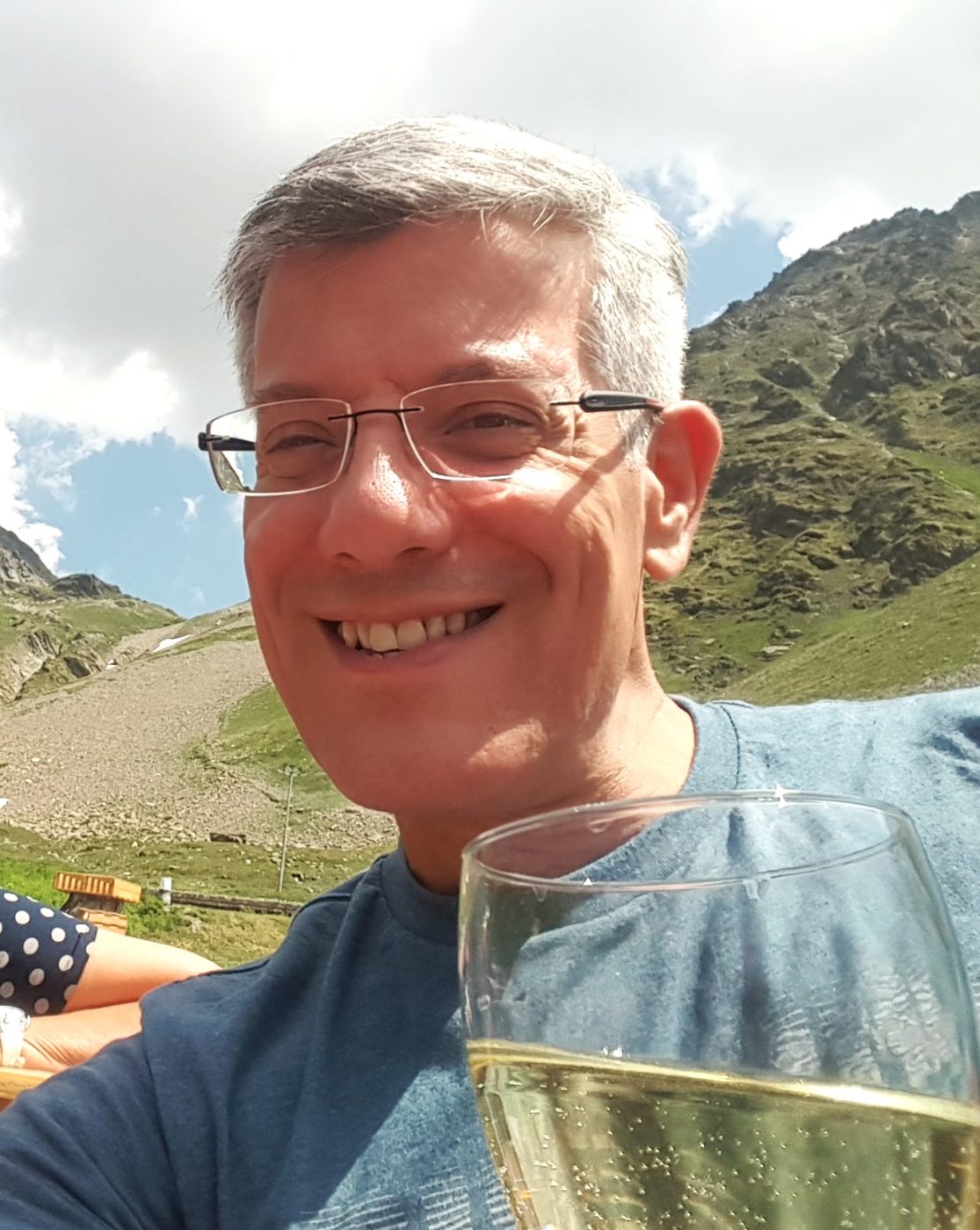 A man with white light hair wearing glasses while outside. He is smiling and holding close to the camera a glass of white wine.
