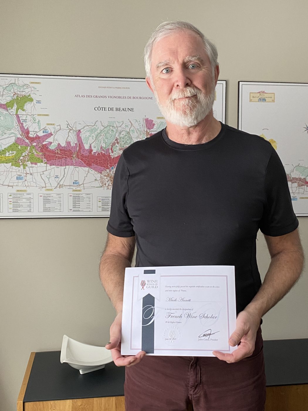 A man with a white beard and white hair wearing a black t-shirt while holding up a French Wine Scholar certificate.