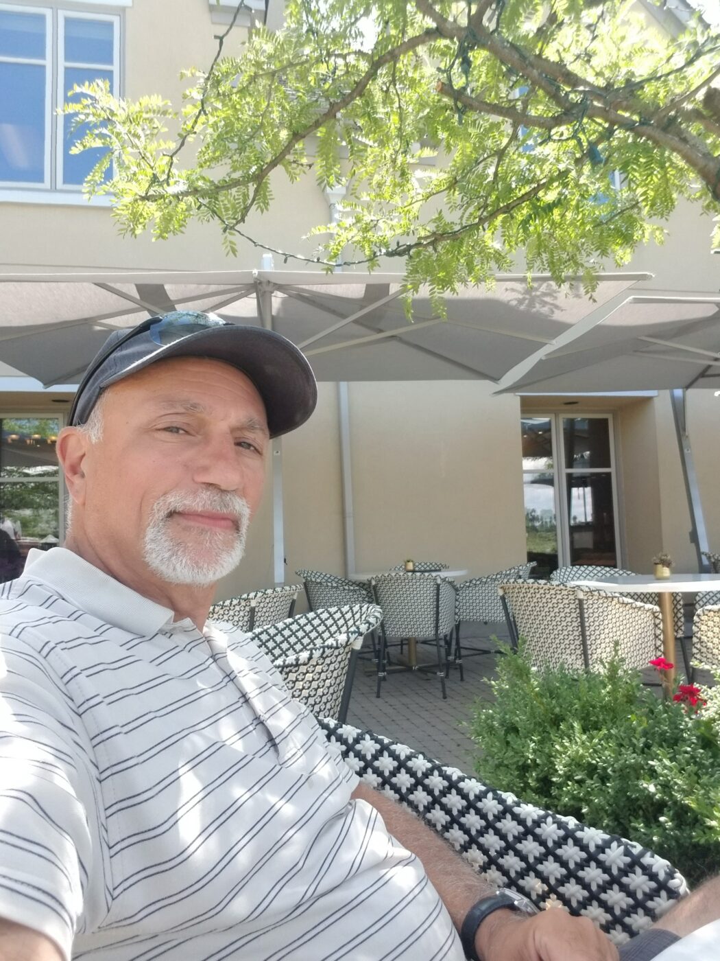 A man sitting outside under a tree near a grey umbrella. He is wearing a grey striped polo shirt and a dary grey ball cap. He has a grey facial hair.