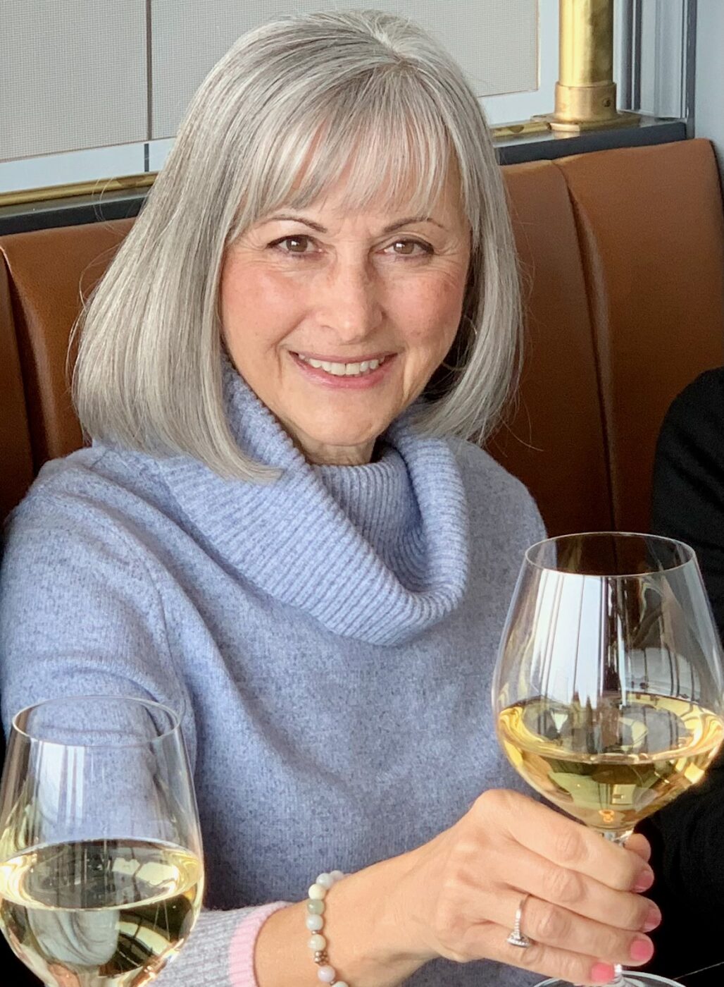 Woman in a light blue cowl neck sweater holding up a glass of white wine. She is smiling and has blue eues and grey hair with bangs.