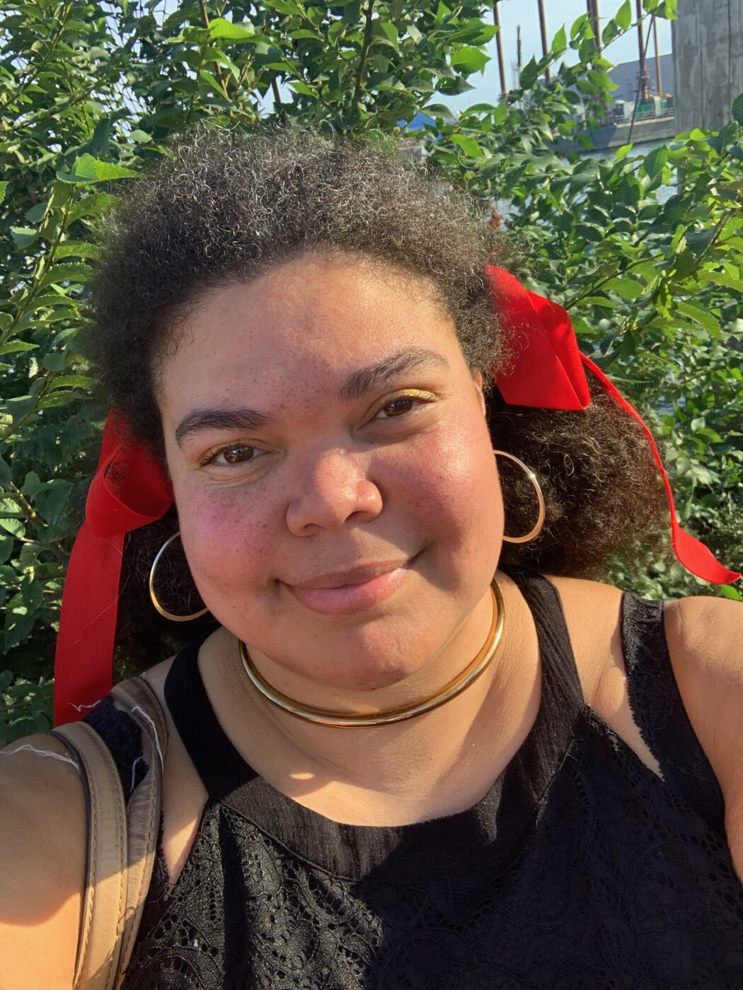 Selfie of a woman outside looking into the camera. She has brown curly hair with a red ribbon tied in the back and big gold hoop earrings.