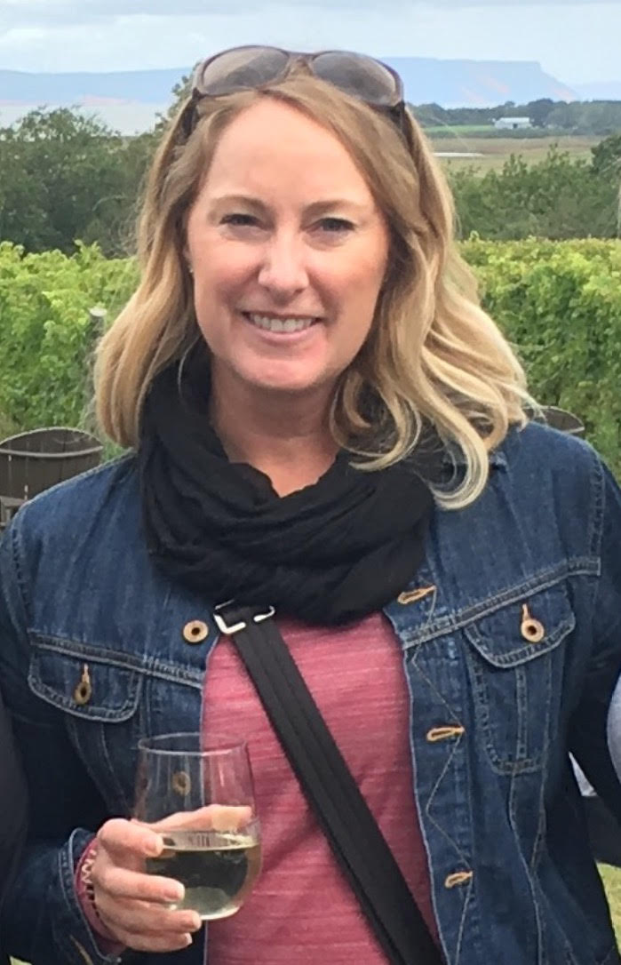 Woman in front of a row of vines in a denim jacket, black scarf and pink shift. She is smiling with sunglasses on her head and a glass of red wine in her hand.