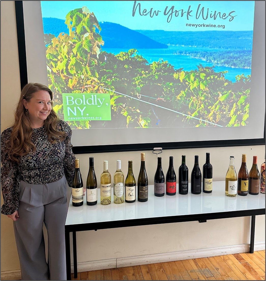 Smiling woman standing by a row of wine bottles on a table with a presentation on New York Wines in the background.