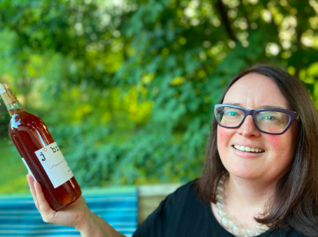 Woman wearing glasses standing outside while holding up a bottle of light red wine.