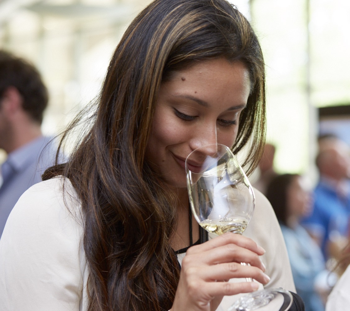 Woman with brown hair in a white shirt leaning into a glass of Chardonnay to smell it.