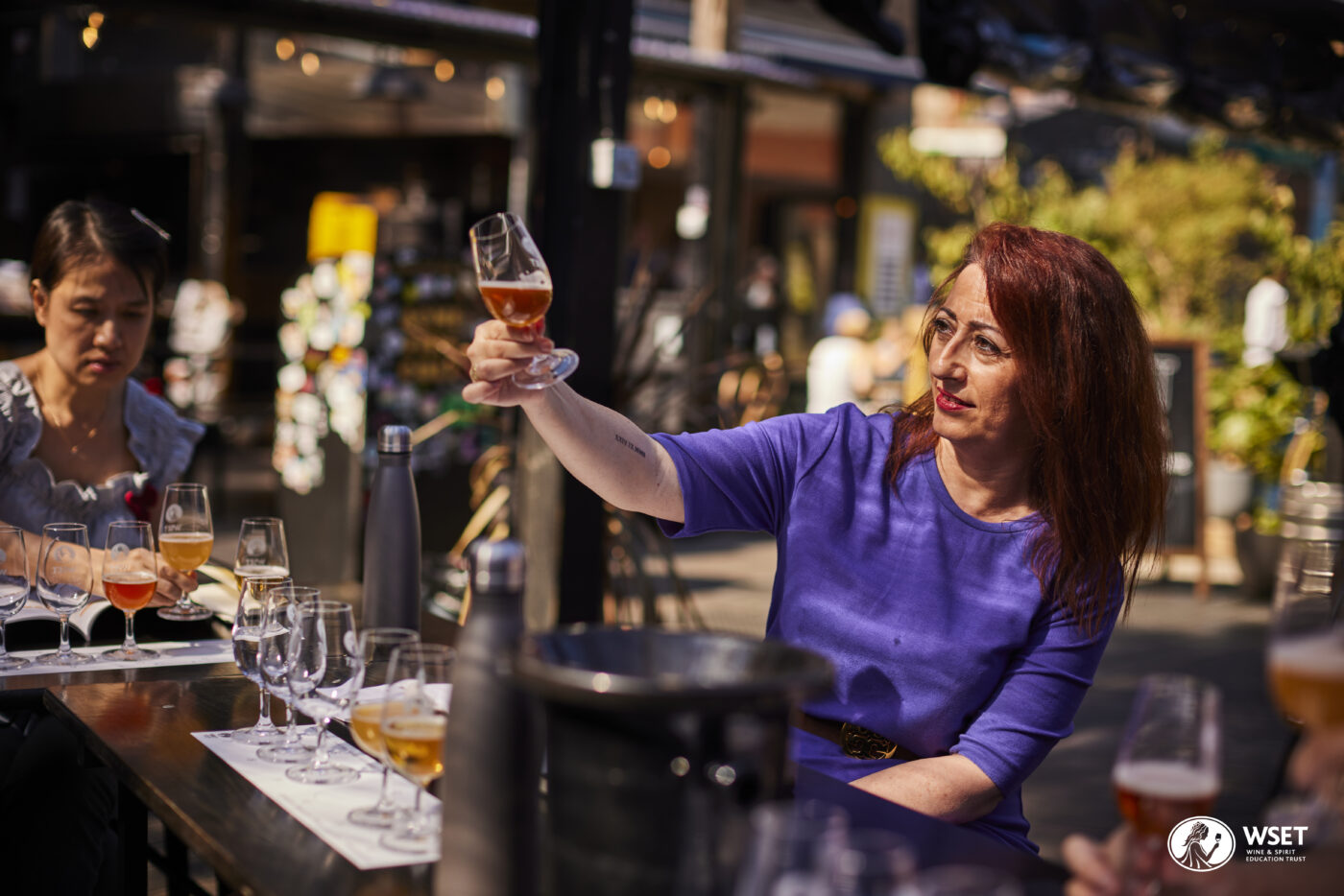 Redheaded woman in a purple shirt behind a bar holding up a beer in a glass.